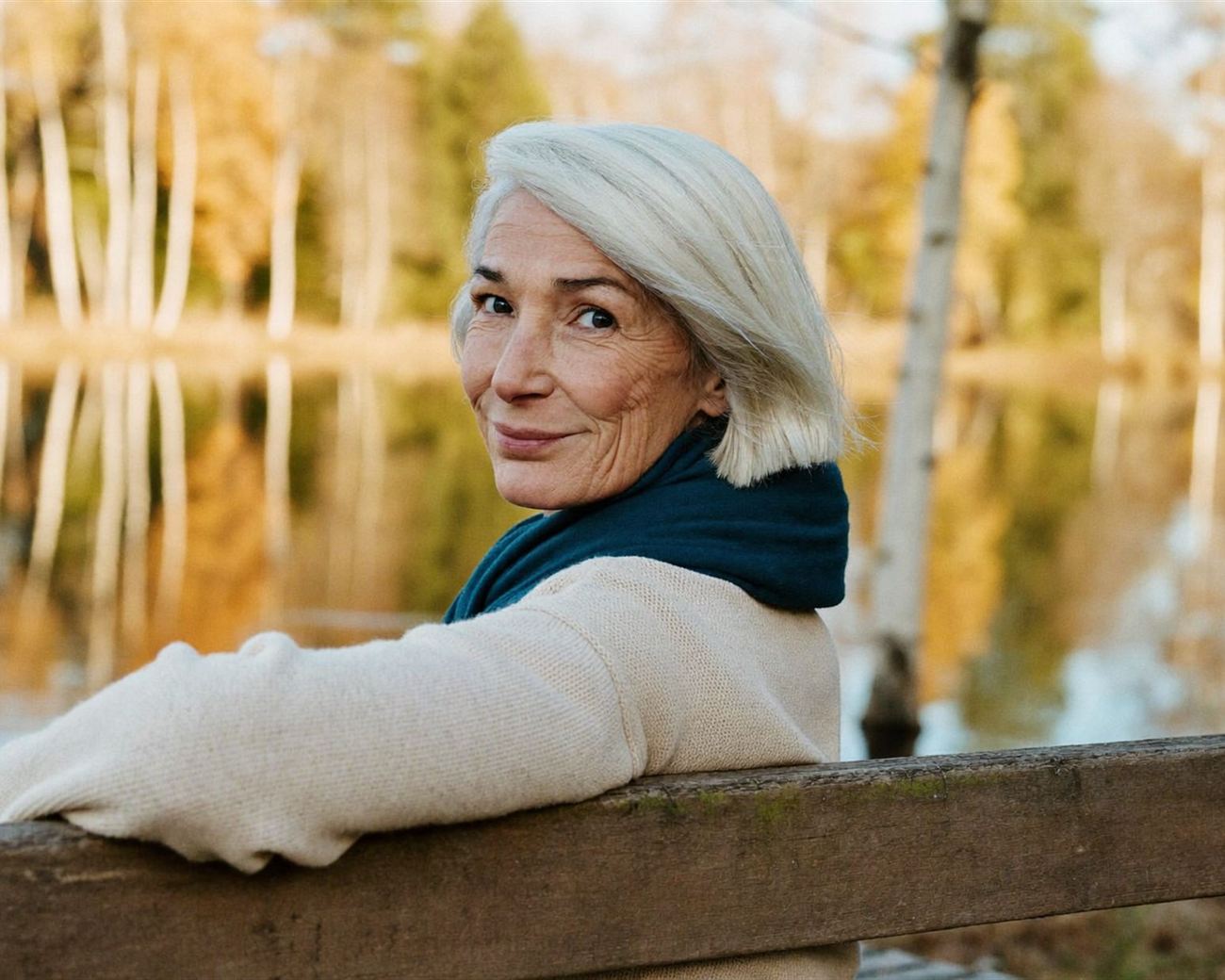 Smilende kvinne med teal skjerf på en benk ved et vann om høsten